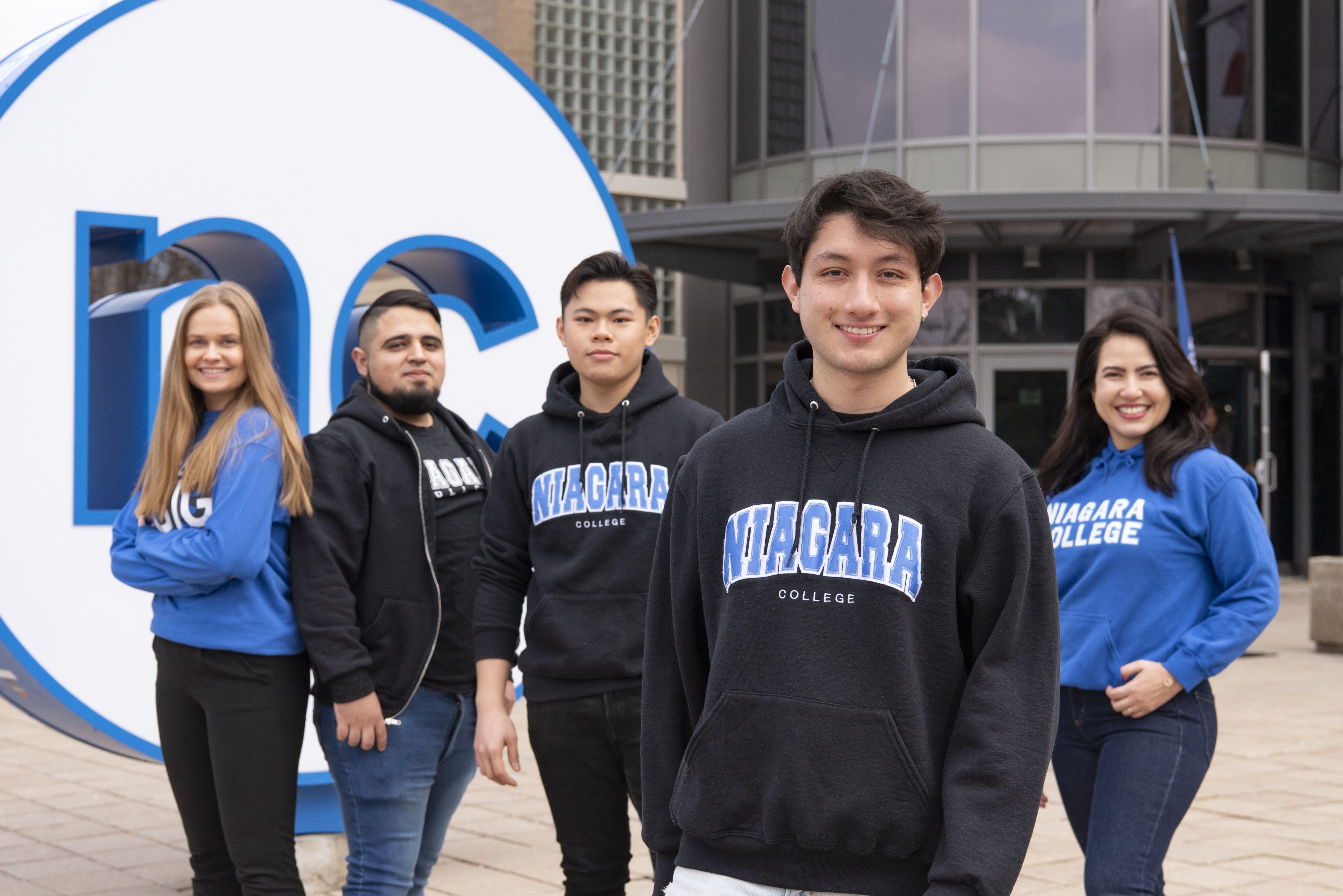 Group of diverse students wearing Niagara College hoodies stands in front of NC button at entrance to DJP campus. 
