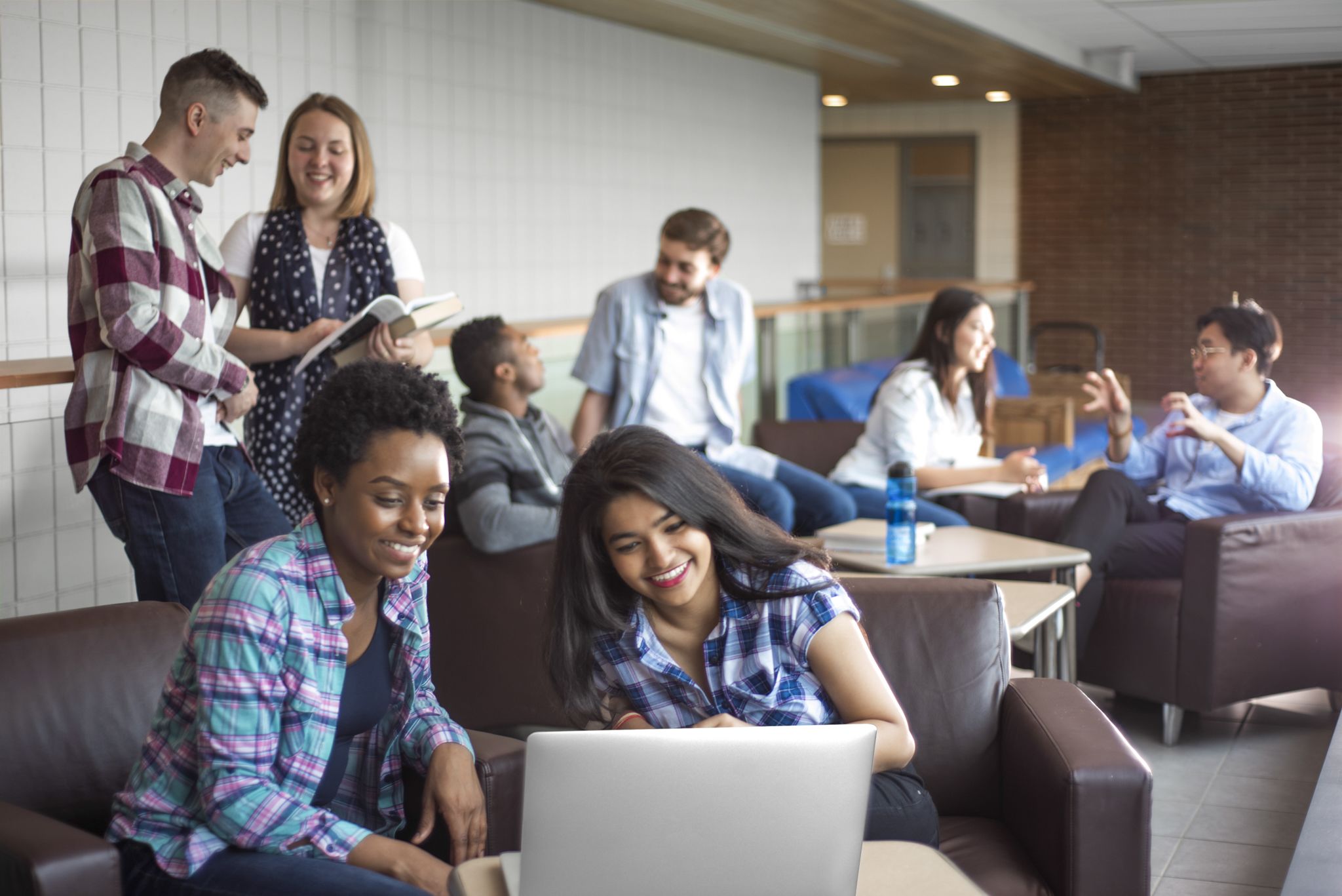 Two seated female students focus on a laptop, surrounded by other students talking in the background.